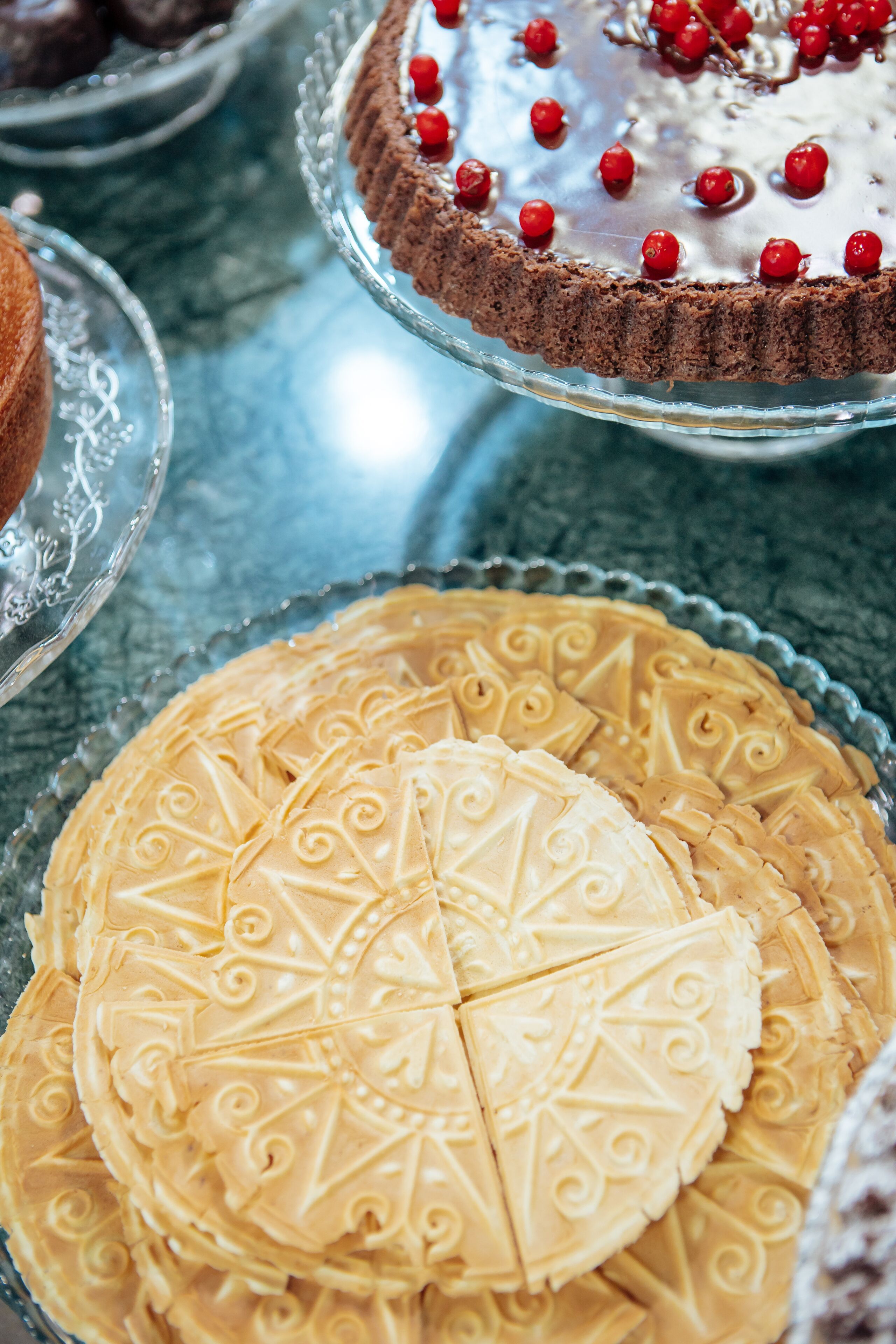A variety of desserts on a glass table, including a chocolate cake with red berries and a white cake with a patterned design.