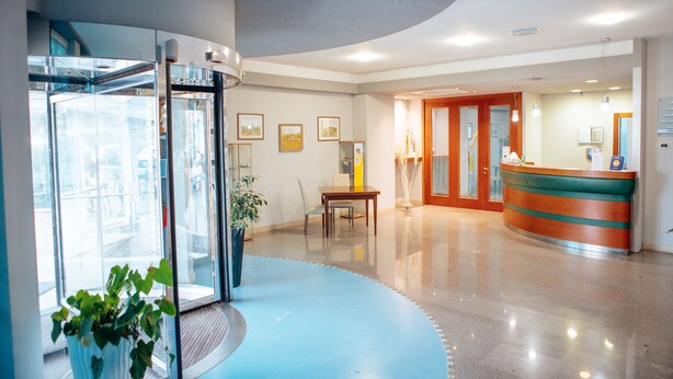 A hotel lobby with a reception desk, a glass elevator, a dining area with a table and chairs, and a potted plant. - Hotel Amico (Citta Sant'Angelo)