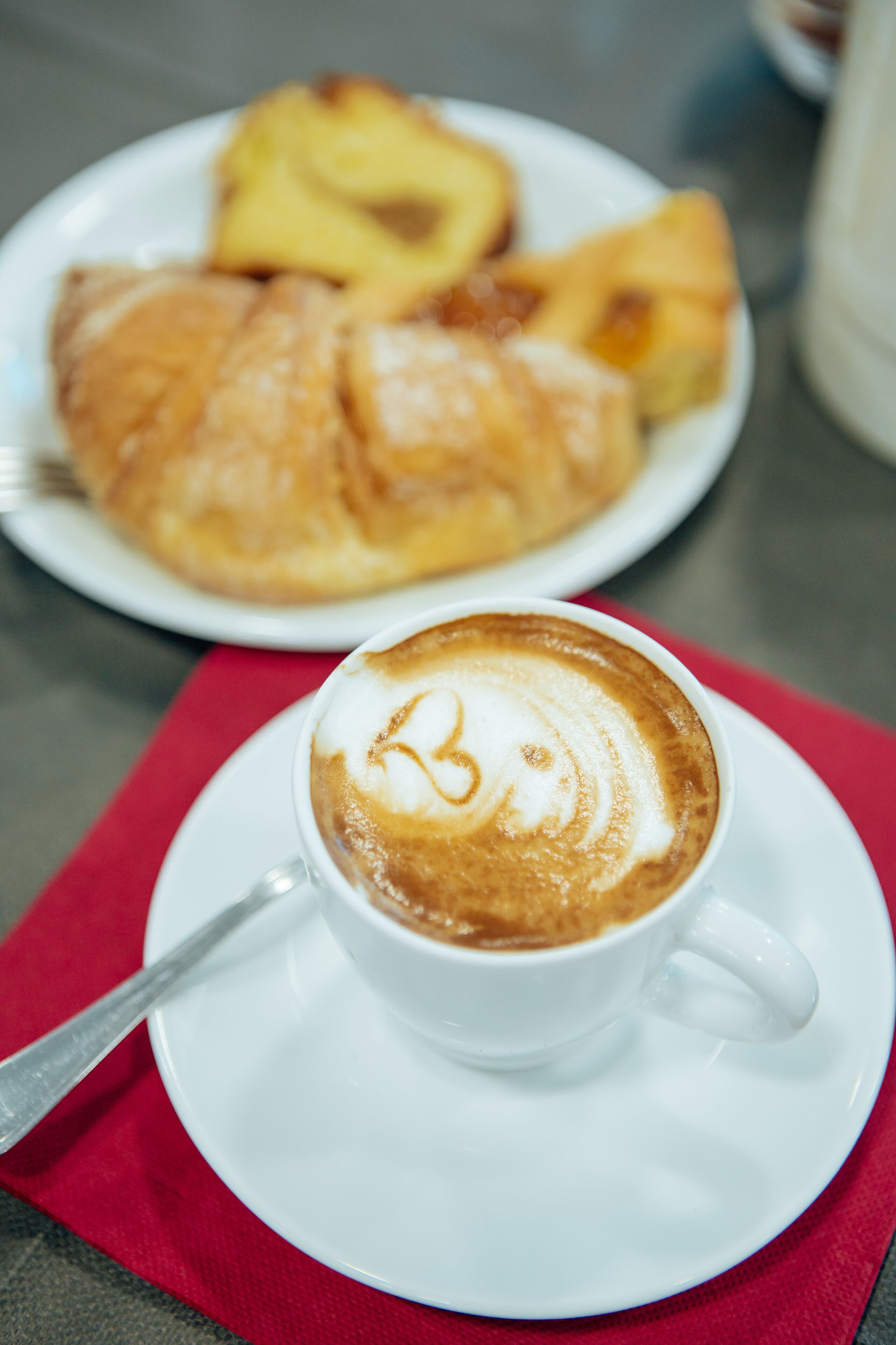 Una taza de café con arte de latte sobre un plato, un plato con croissants y un mantel individual rojo sobre una mesa.