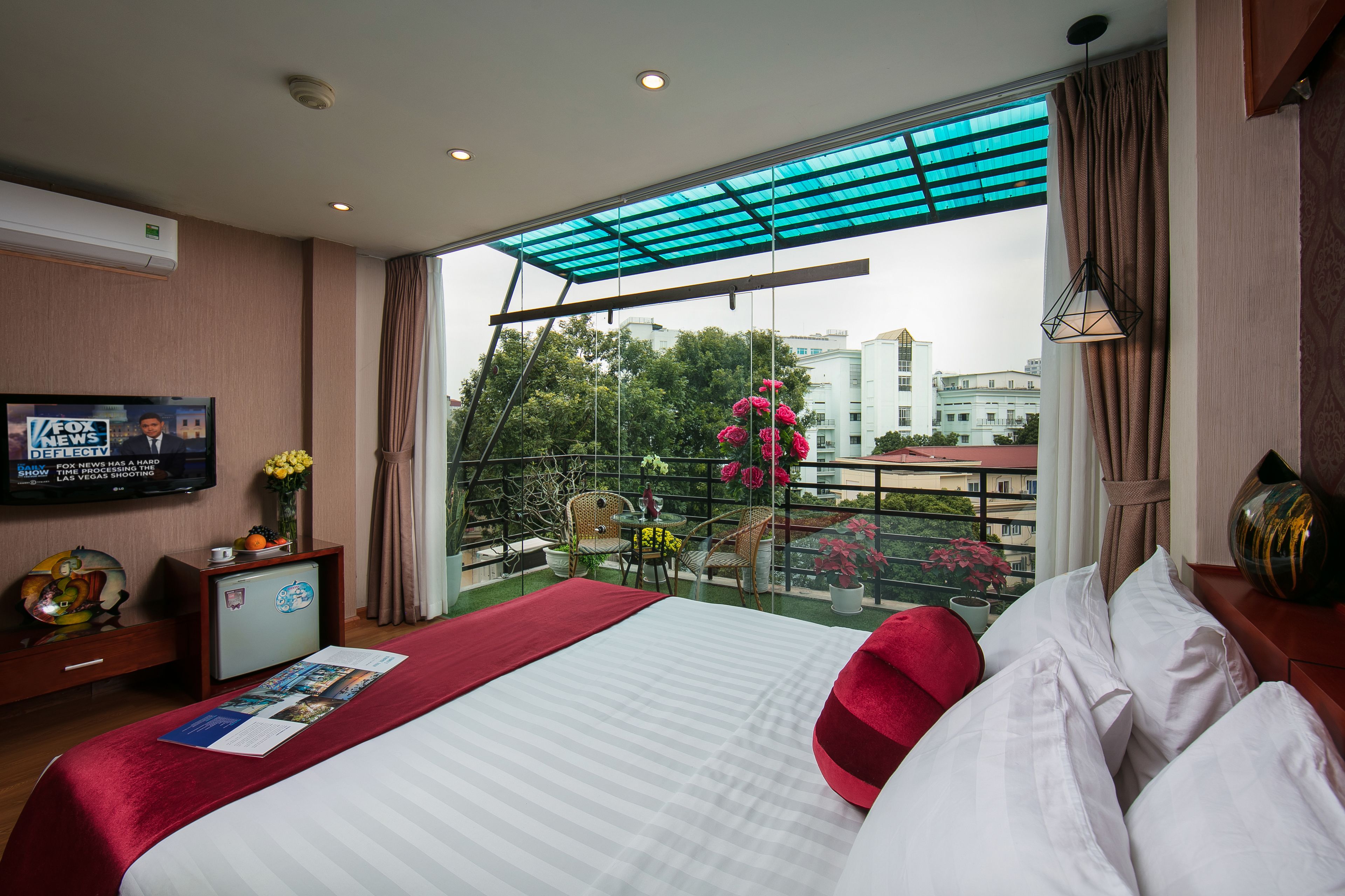 A hotel room with a bed, a TV, a balcony with a view of buildings and trees, and a small table with chairs.