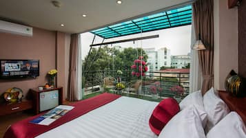Een hotelkamer met een bed, een televisie, een balkon met uitzicht op gebouwen en bomen, en een kleine tafel met stoelen.