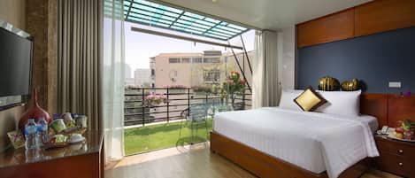 A hotel room with a large bed, a TV, a desk with a vase and bottles, a balcony with a view of buildings and flowers.