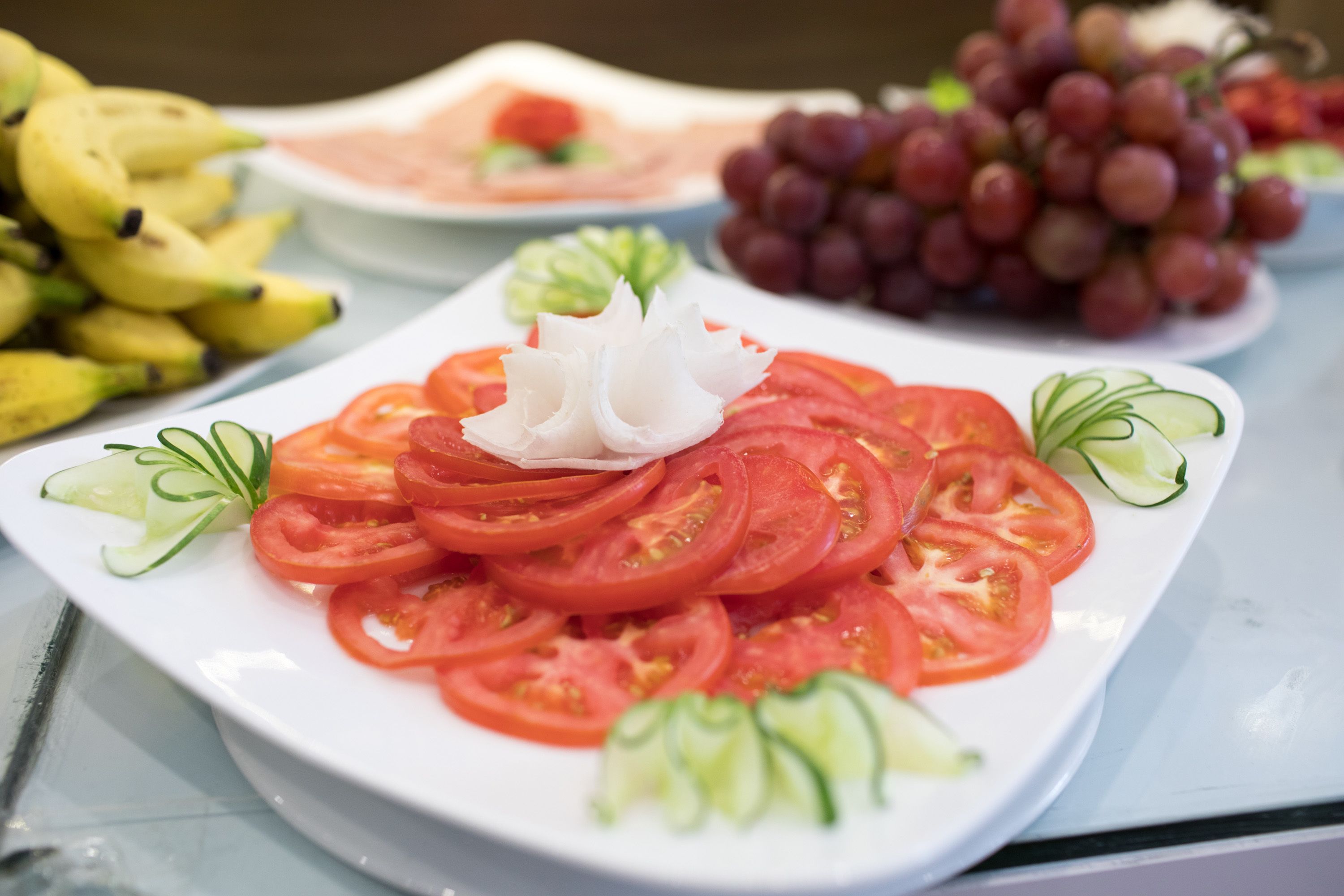 A plate of sliced tomatoes with cucumber and green onion garnish, bananas, and grapes on a table.