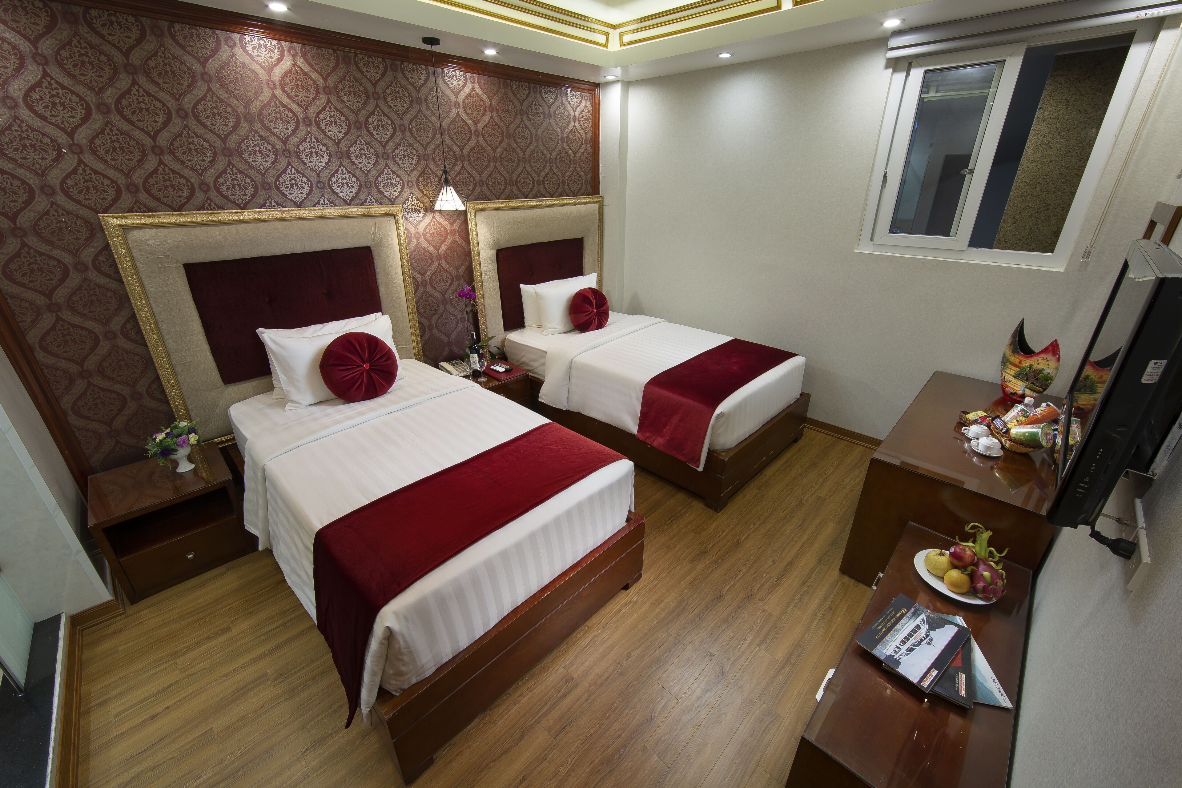 A hotel room with two beds, wooden flooring, a desk with a fruit bowl, and a window.