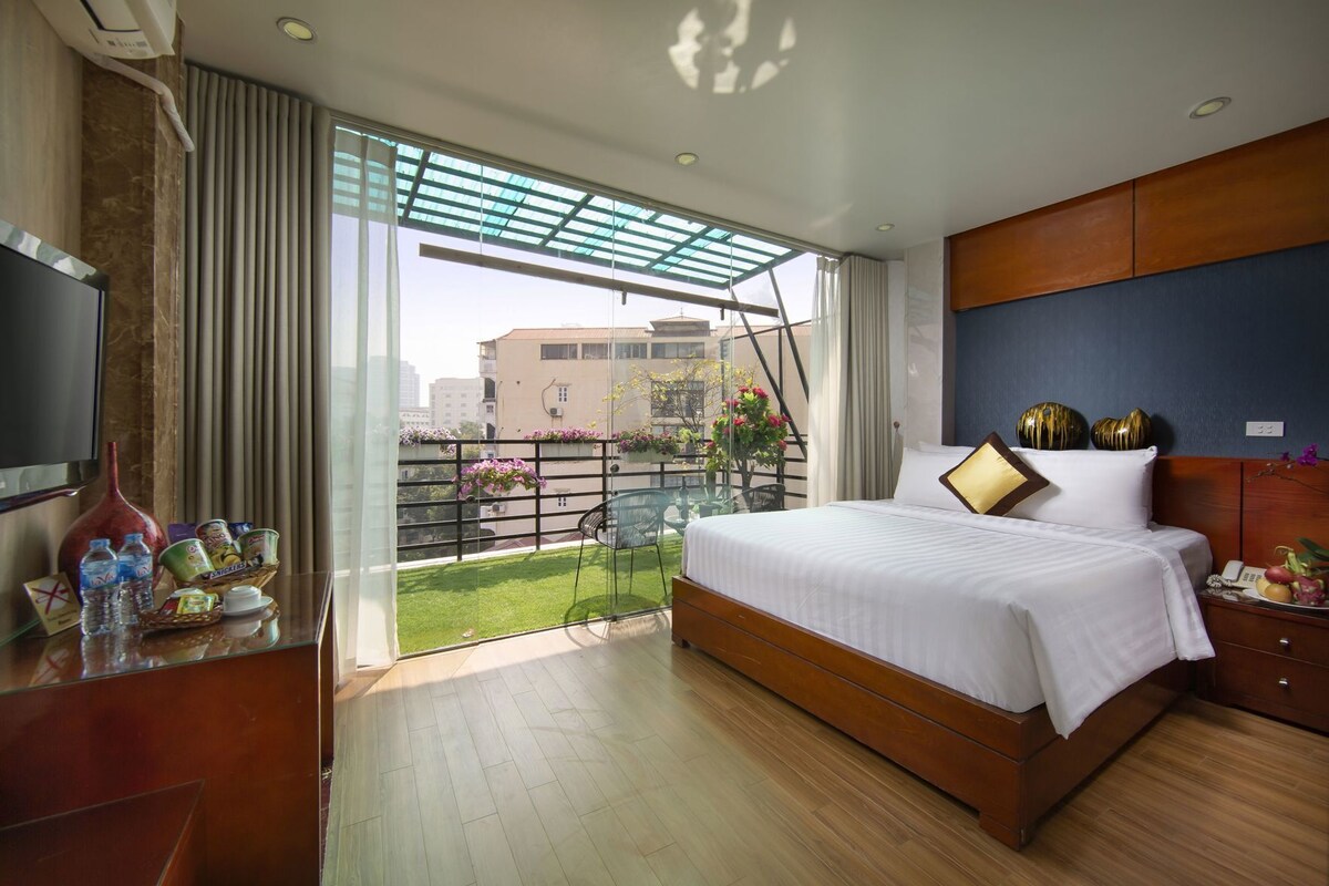 A bedroom with a bed, a TV, a desk with toiletries, and a balcony with plants.
