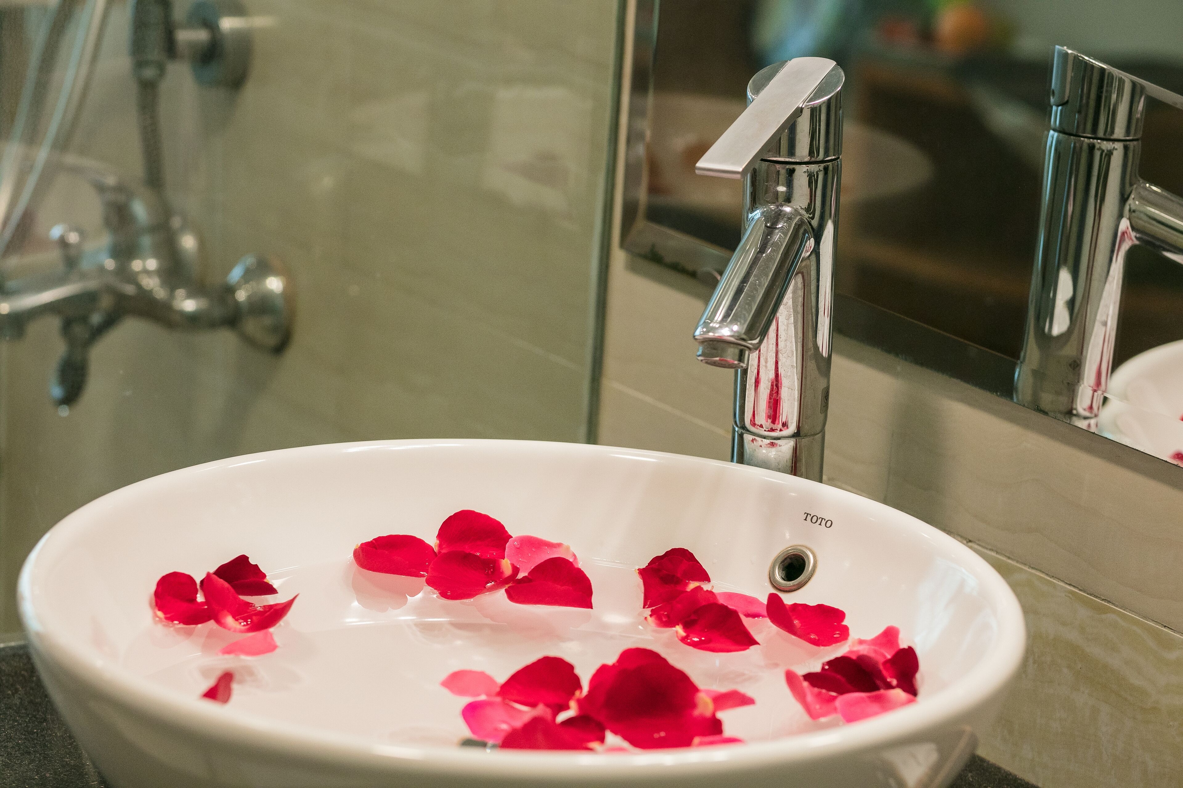 A white bathroom sink with rose petals floating in the water, a chrome faucet, and a mirror reflecting the sink and faucet.