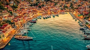Uma vila costeira com edifícios à beira-mar, um porto com barcos e uma paisagem montanhosa ao fundo.