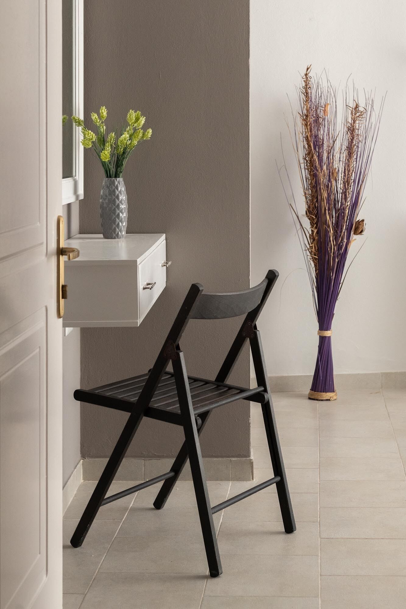 Un baño moderno con un tocador blanco, una silla negra y un jarrón con plantas secas.