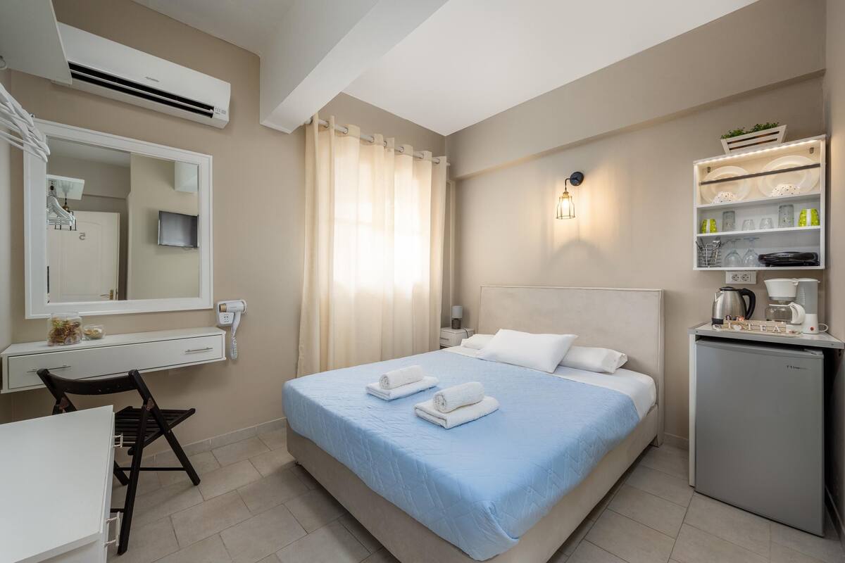 Quarto de hotel com cama, escrivaninha, cadeira, mini-geladeira e prateleira com artigos de higiene pessoal.