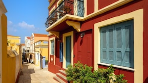 Uma rua estreita com edifícios coloridos, uma placa visível com 'MELISSA'S ROOMS' e degraus que levam a uma porta.