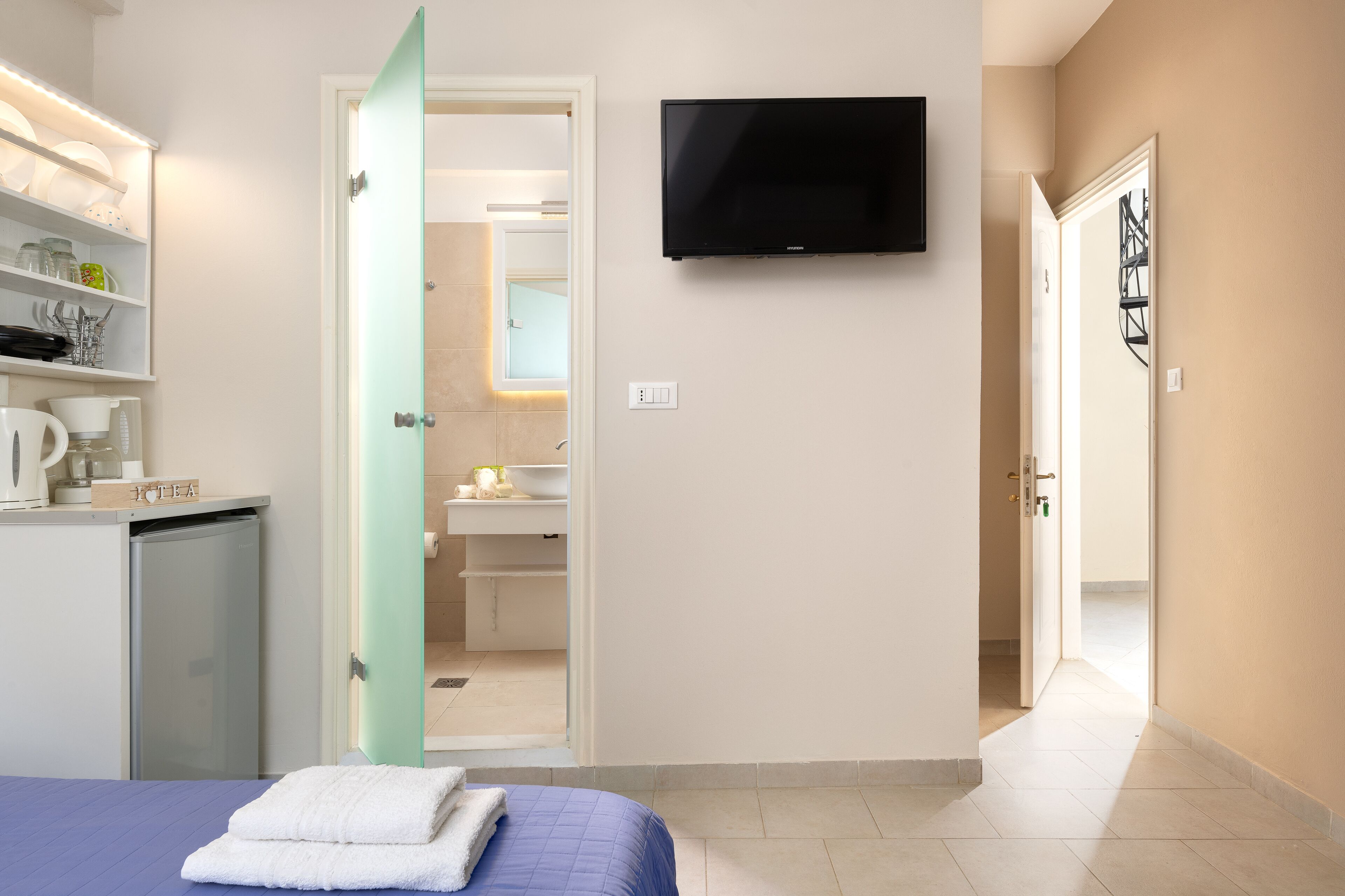 Habitació d'hotel amb un llit, una televisió penjada a la paret, una cuineta i un bany visible a través d'una porta oberta.