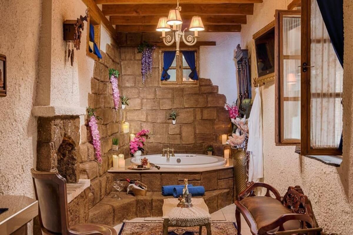 A stone-walled bathroom with a bathtub, wooden chairs, and a chandelier.