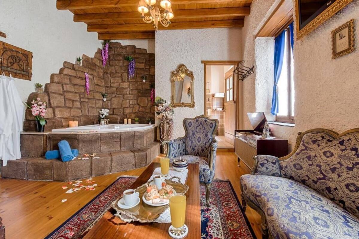 A cozy room with a stone wall, a wooden ceiling, a patterned sofa, a dining table with a tray of food and drinks, and a chair with a decorative mirror.