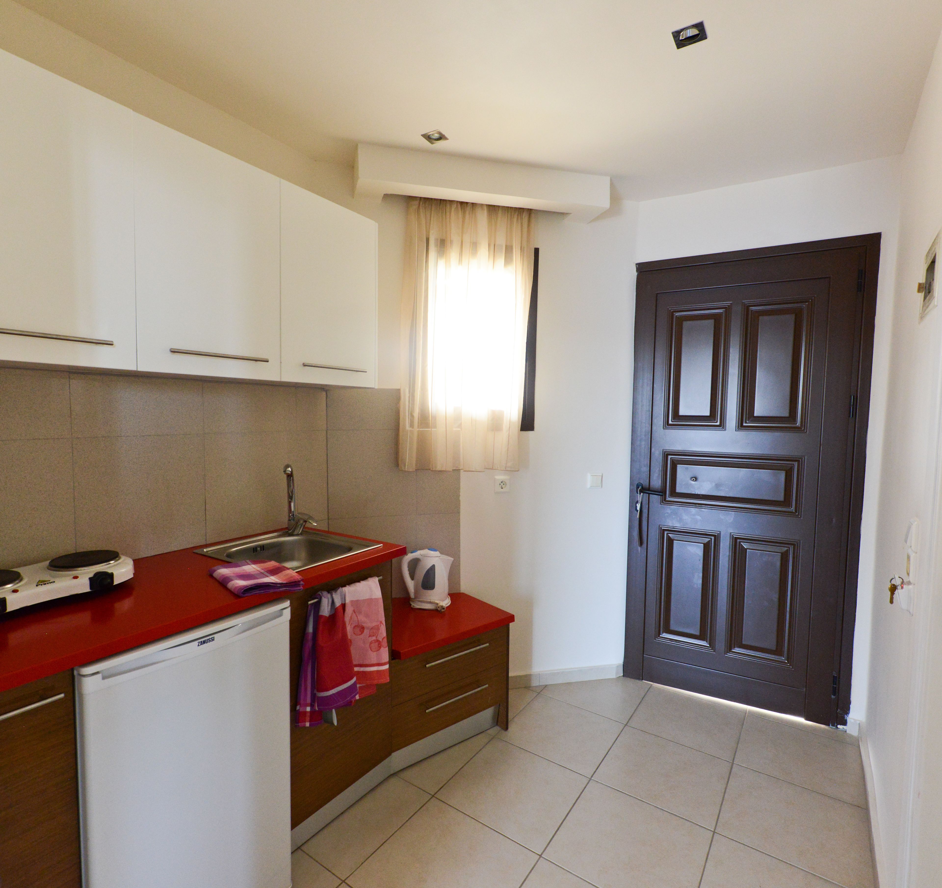 A compact kitchen with white cabinets, a red countertop, a small refrigerator, a kettle, and a dark wooden door.