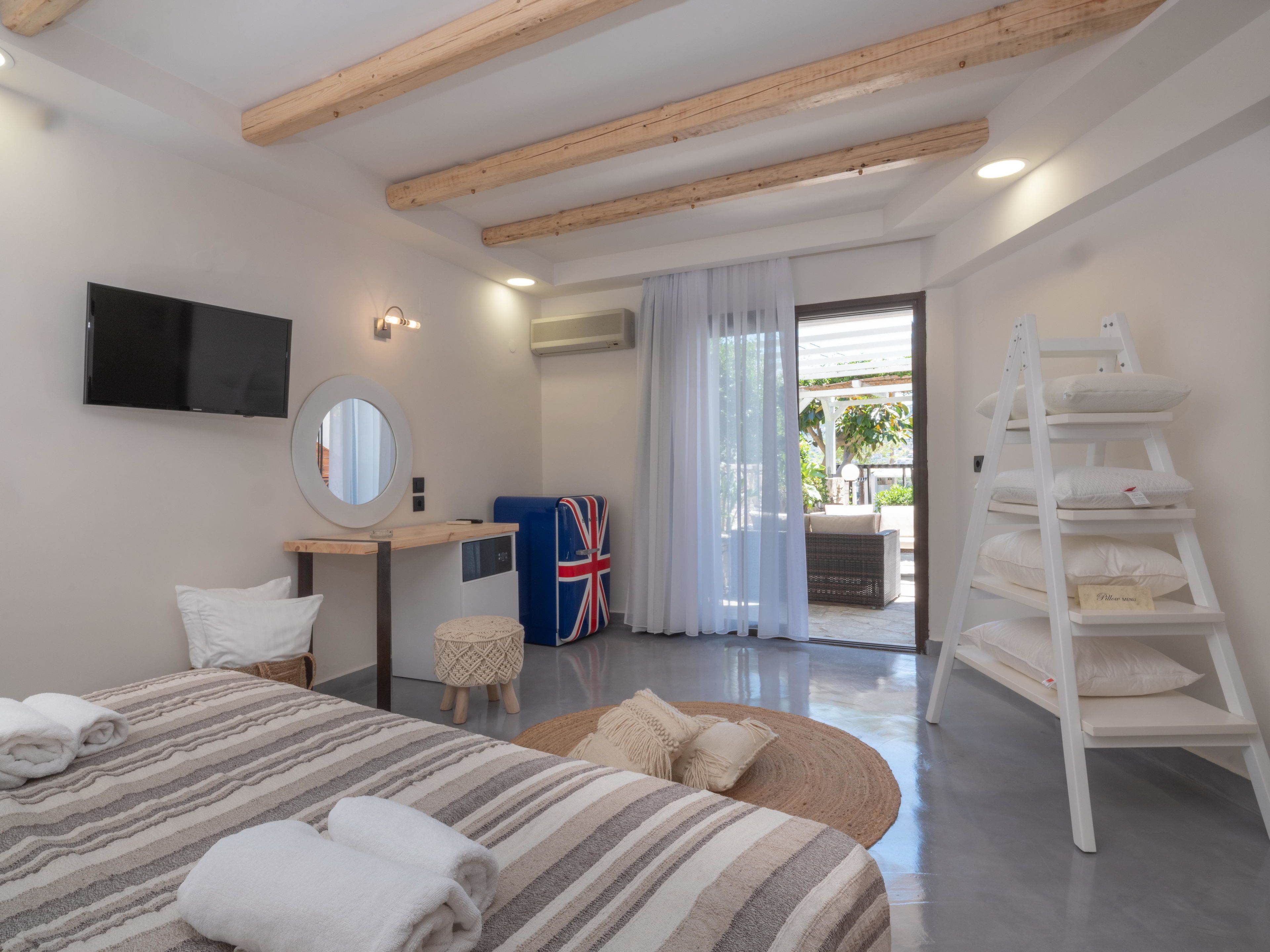 A modern bedroom with a bed, a TV, a mirror, a desk, a chair, a small stool, a shelf with pillows, and a balcony with a view.