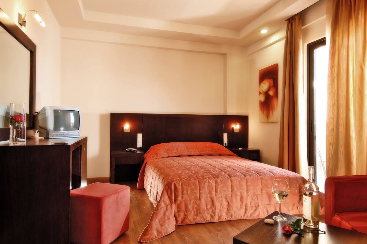 Une chambre d’hôtel avec un lit, une télévision, une table de chevet, une petite table, une chaise rouge, une table en bois avec un verre à vin et une bouteille, et un tableau accroché au mur.