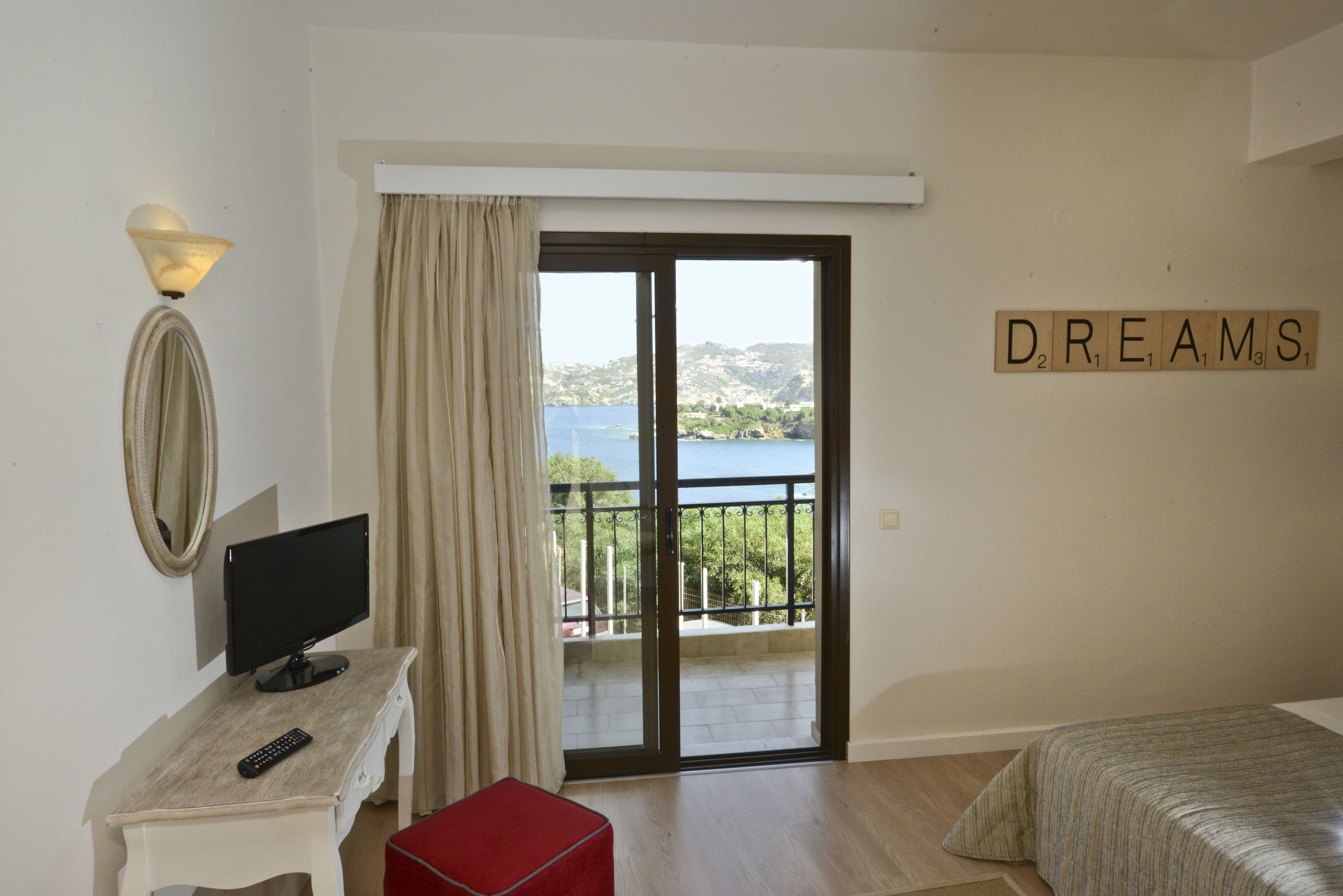 A bedroom with a bed, a desk with a computer monitor, a red ottoman, a window with a view of the sea, and a wall with the word "DREAMS" spelled out in tiles.