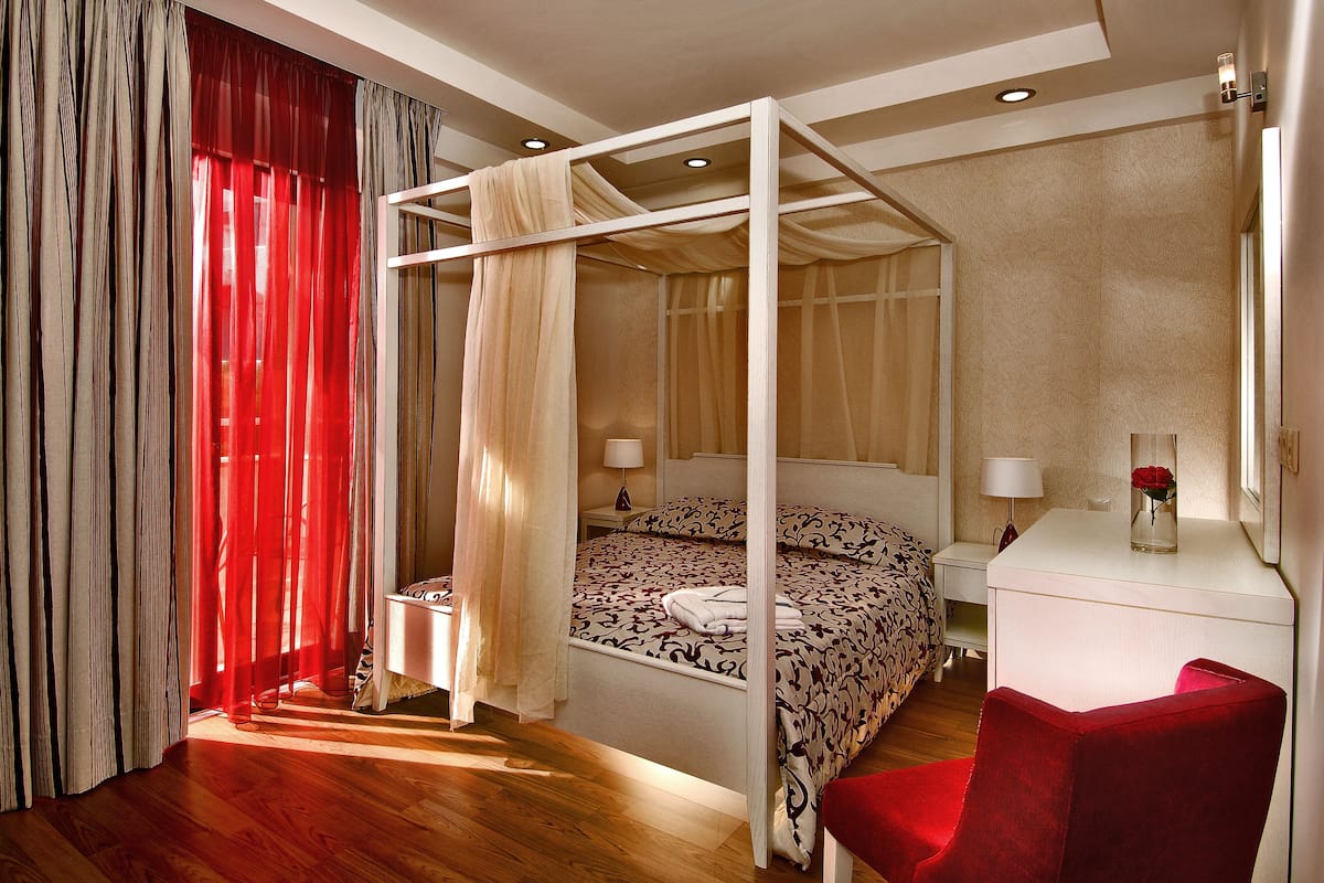 Un lit à baldaquin à quatre poteaux, avec une table de chevet et une lampe, une chaise rouge et un vase avec une fleur sur un bureau.