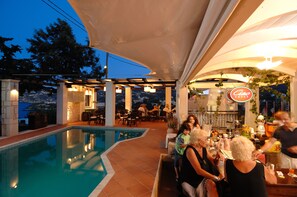 Ein Poolbereich im Freien mit überdachter Terrasse, Esstischen und Blick auf die Stadt.
