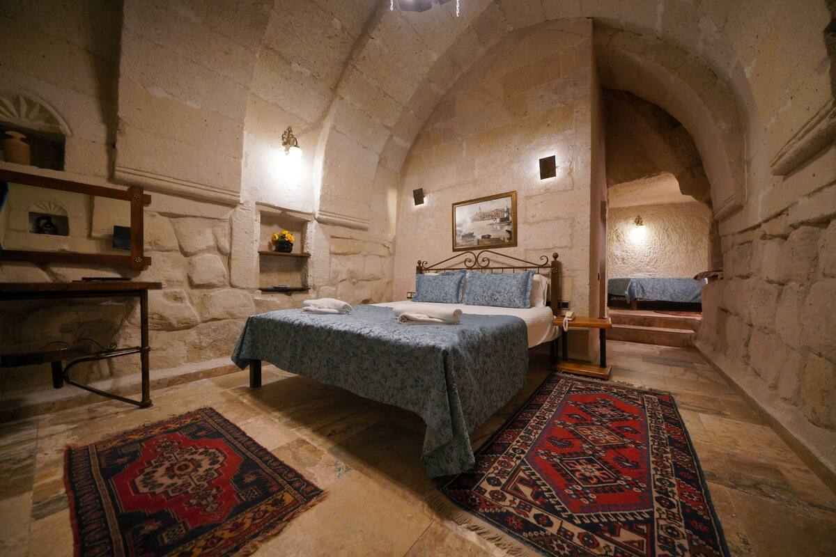 A stone-walled bedroom with a bed, a nightstand, a wooden table, and a patterned rug.