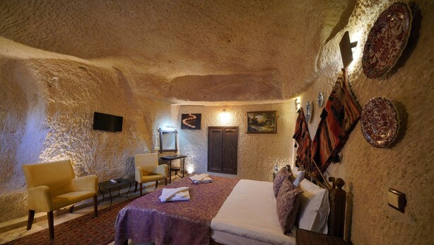 A bedroom with a bed, two chairs, a small table, and a wall-mounted TV. - Emit Sakura Cave Hotel (Nevsehir)