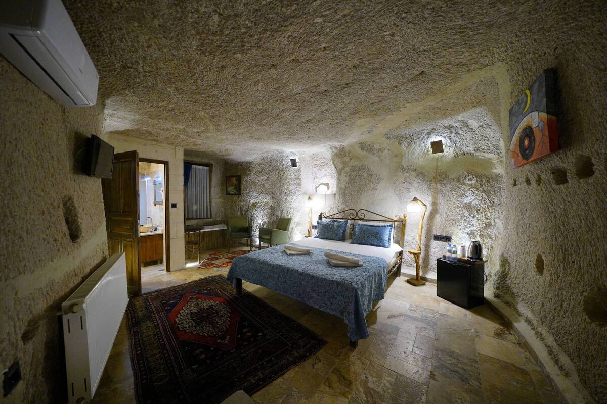 A bedroom with a stone ceiling, a bed with a blue bedspread, a bedside table with a lamp, and a radiator.