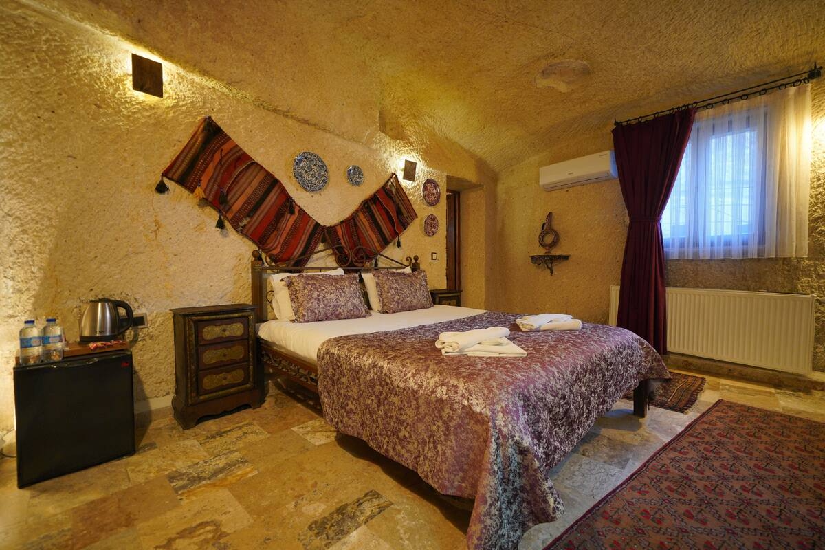 A bedroom with a large bed, a small bedside table, a radiator, and a window with curtains.