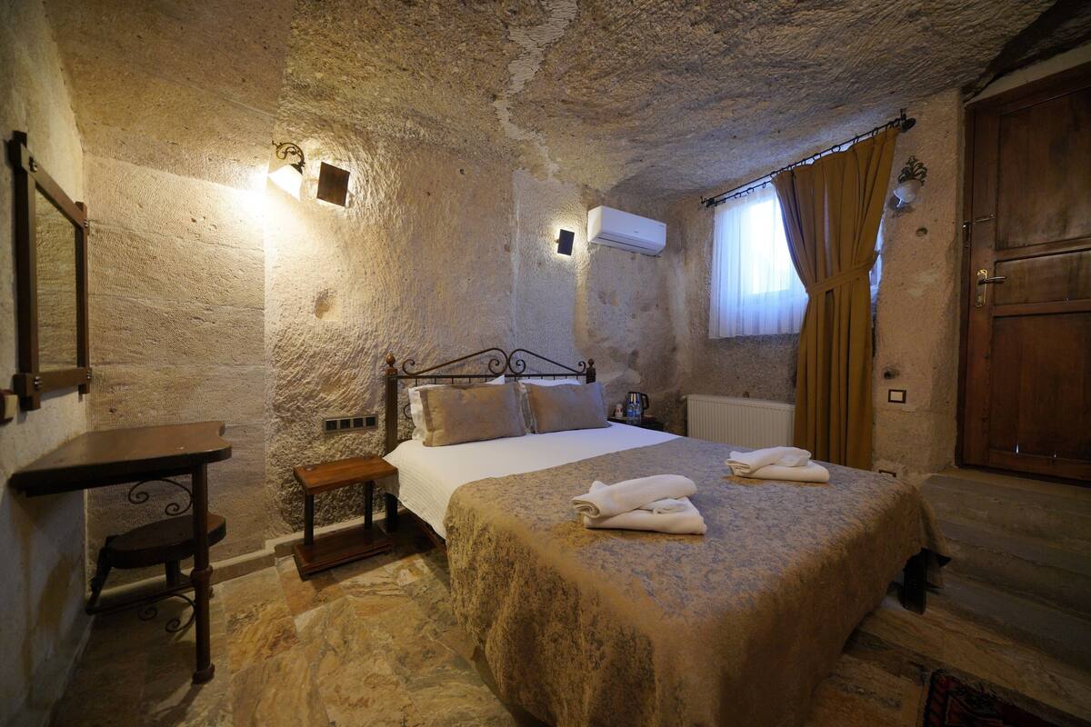 A bedroom with a stone wall, a bed with a blanket, a bedside table, a nightstand, and a wooden door.