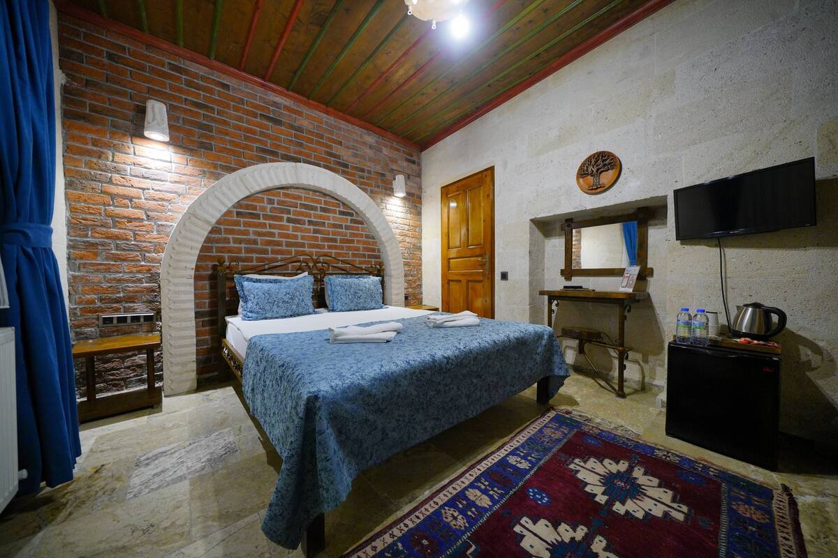 A bedroom with a brick wall, a wooden door, a bed with blue bedding, a TV, and a small table with a kettle.