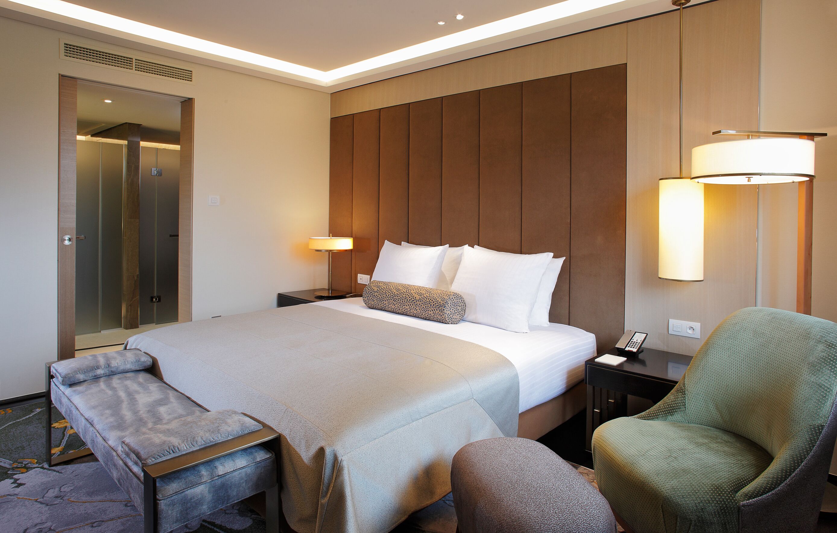A hotel room with a large bed, a bench, a chair, a bedside table with a lamp, and a mirror.