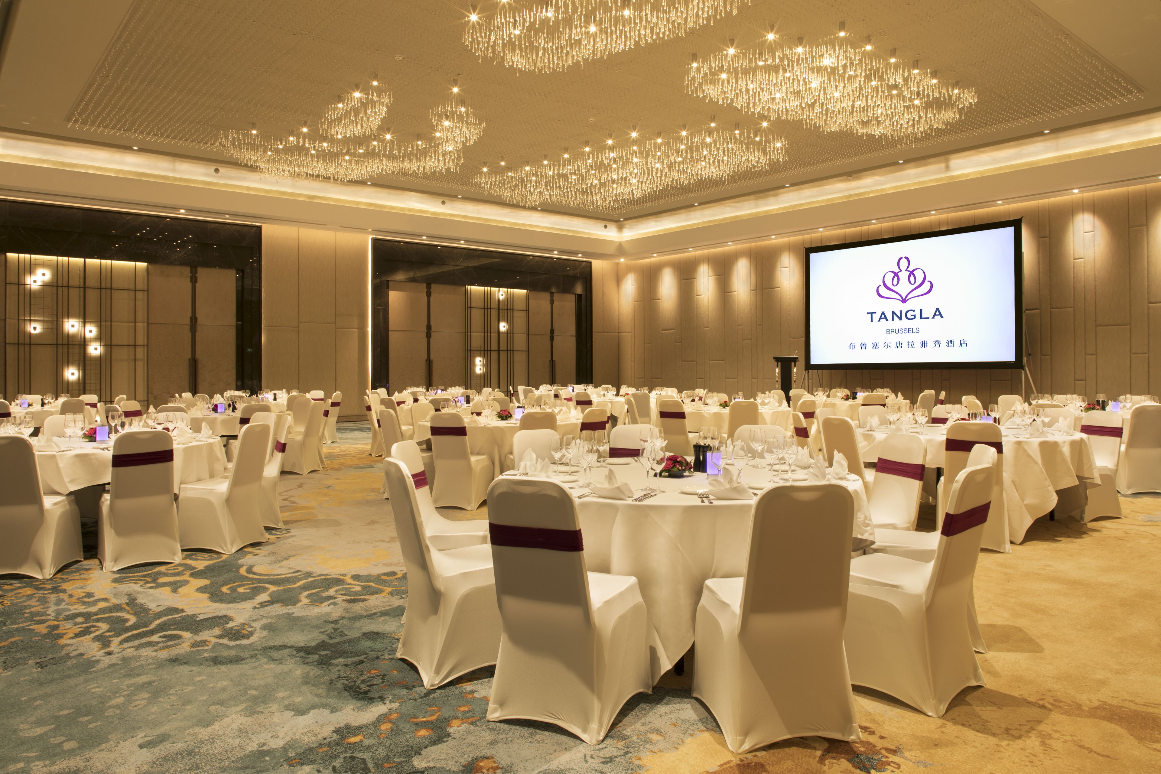 A banquet hall with round tables set for a formal event, featuring a large screen displaying the Tangla Brussels logo.