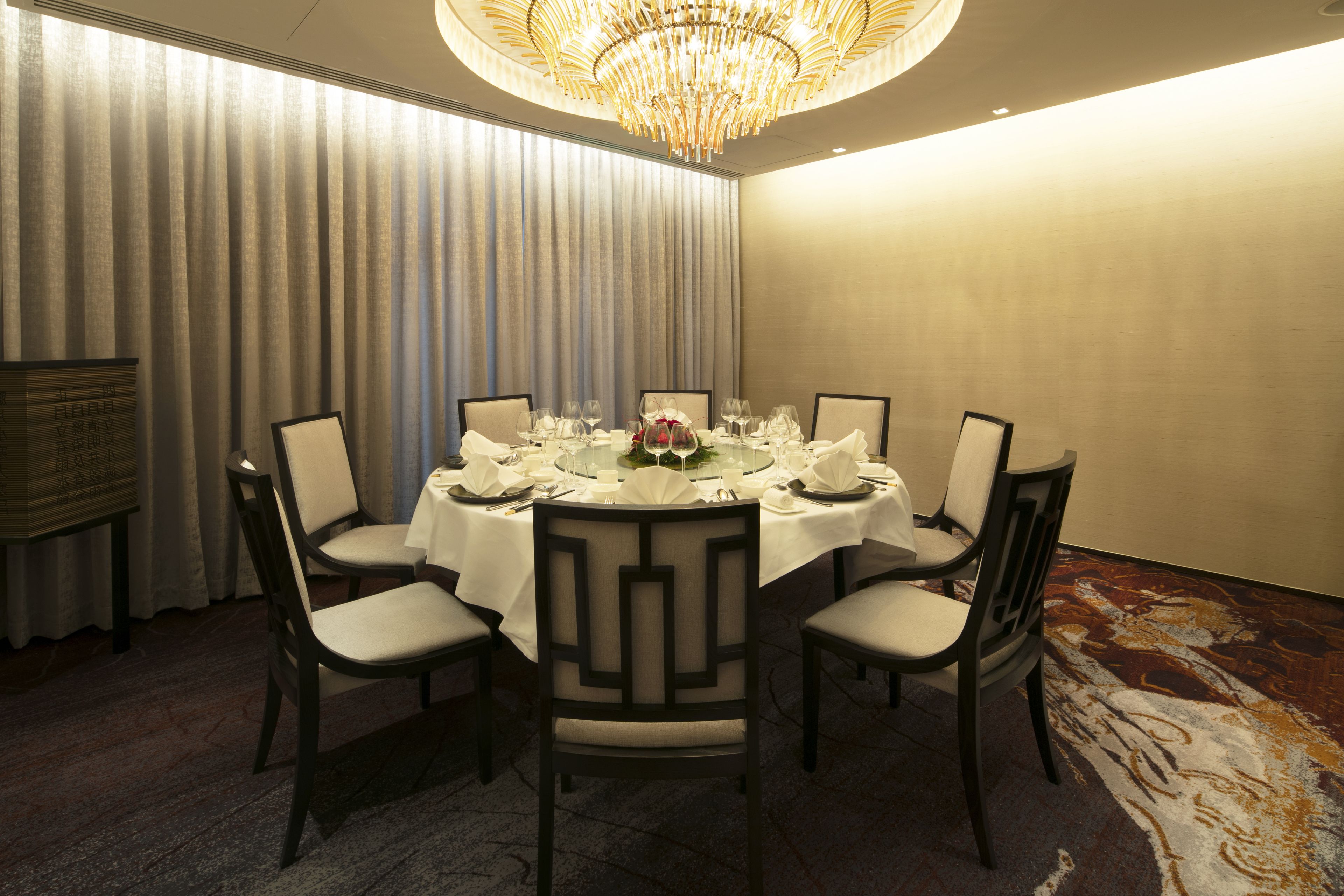 A dining room with a round table set for six, surrounded by chairs, a chandelier, and a large window with curtains.