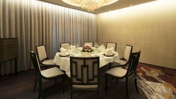 A dining room with a round table set for six, surrounded by chairs, a chandelier, and a large window with curtains.