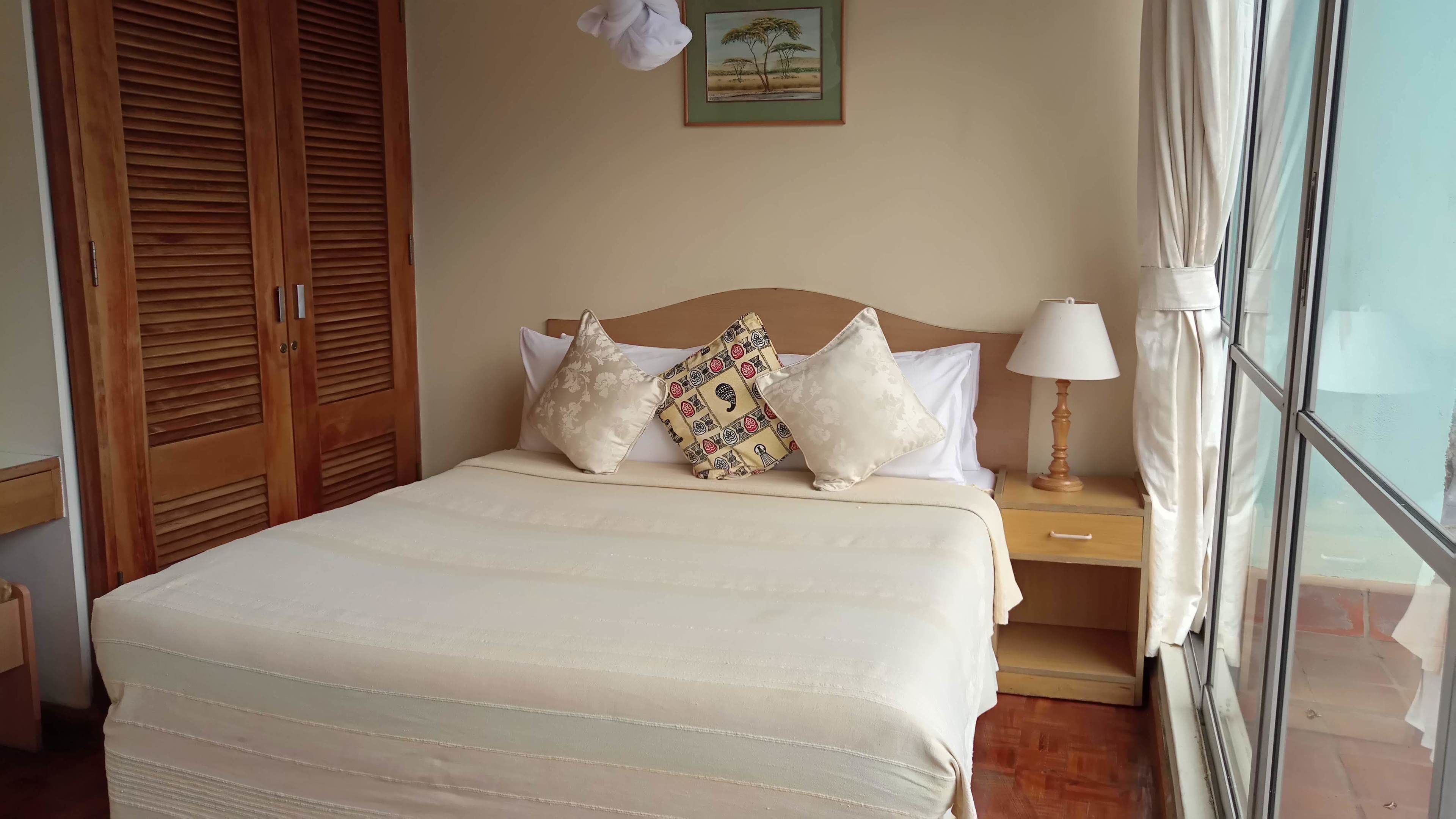 Una cama bien hecha con un cojín estampado, un cabecero de madera, una mesita de noche con lámpara y una ventana con cortinas.