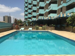 Una piscina en la azotea con sillones de descanso y una zona de estar techada, rodeada por un edificio de gran altura.