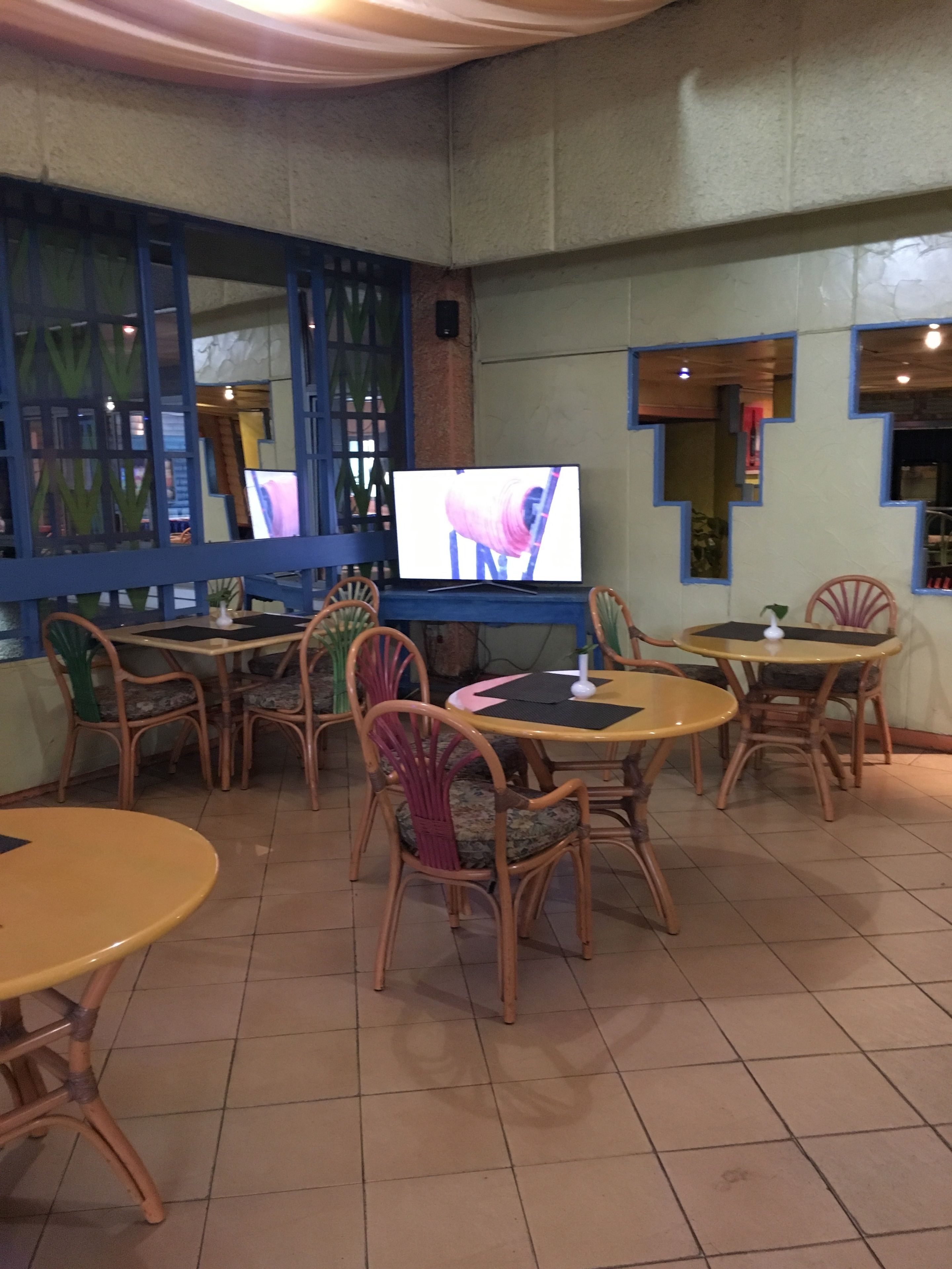 A restaurant interior with round tables and wicker chairs, a television mounted on the wall, and blue-framed windows.