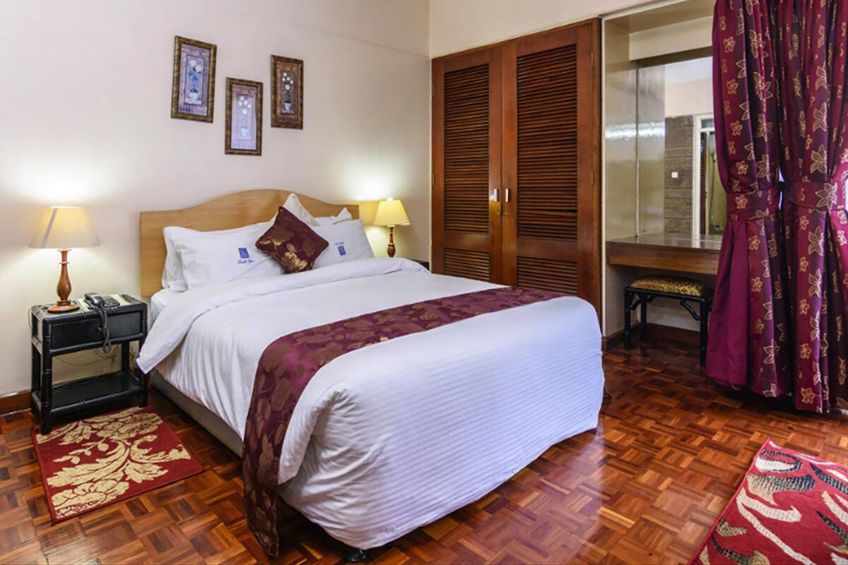 Habitación de hotel con una cama grande, dos lámparas de noche, un armario de madera, un pequeño banco y una alfombra roja con diseño.