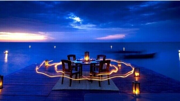 Une table dressée pour un dîner paisible sur une terrasse en bois, avec vue sur l’océan et un bateau à l’horizon.