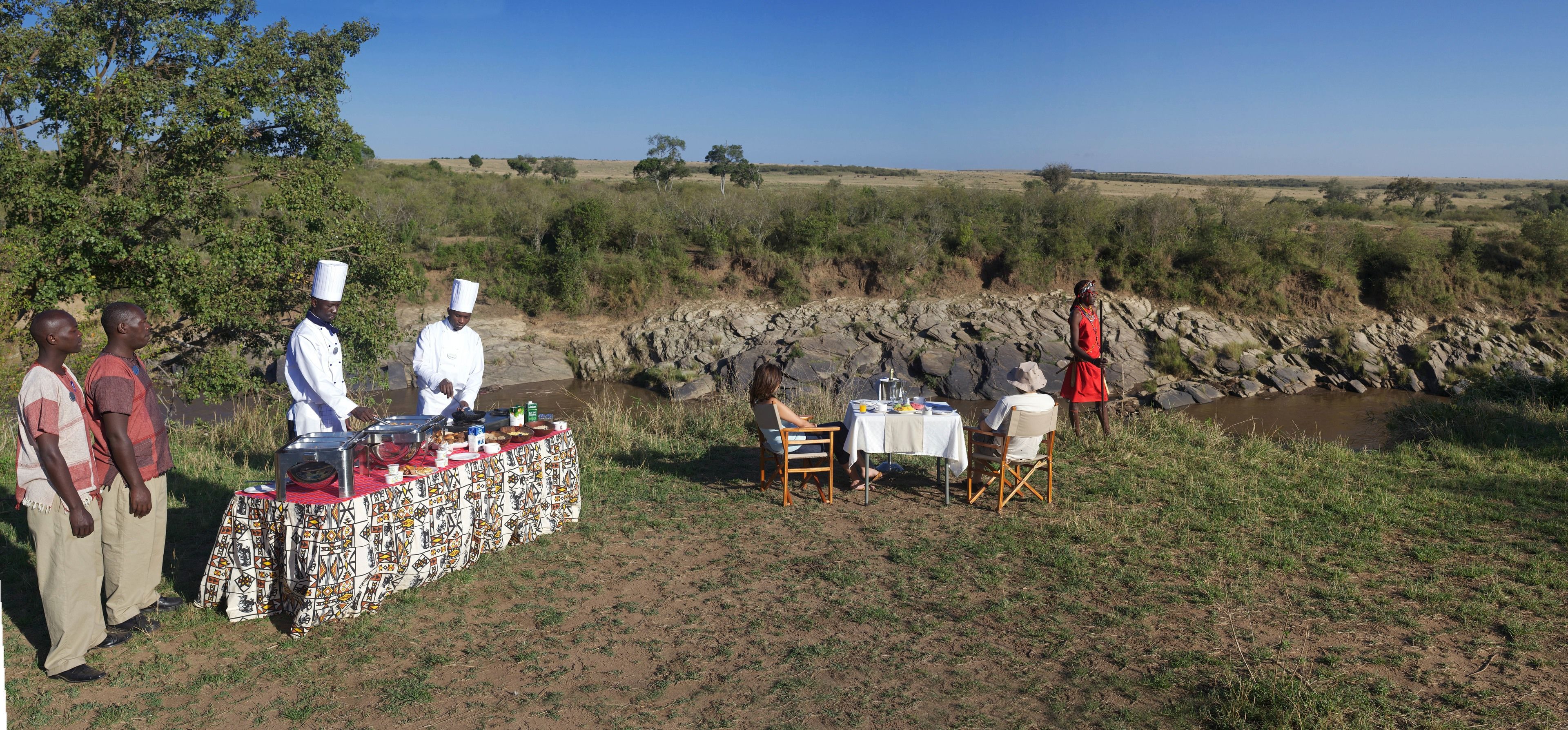 Un grupo de personas al aire libre junto a una mesa preparada para una comida, una orilla de río rocosa y vegetación escasa al fondo.