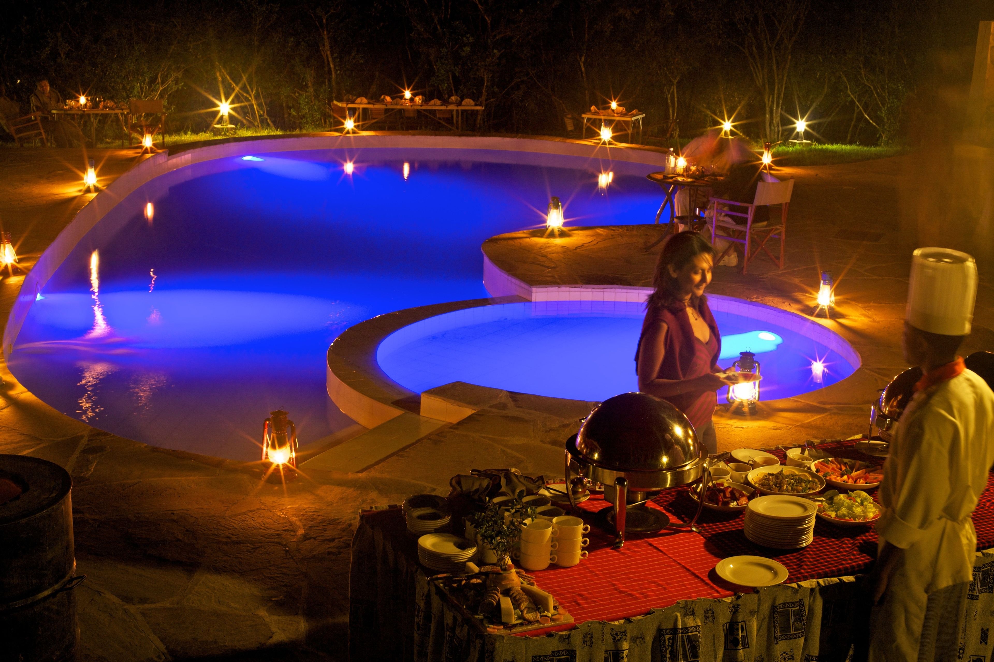 A poolside dining area with a table set for a meal, surrounded by candles and a chef in uniform.