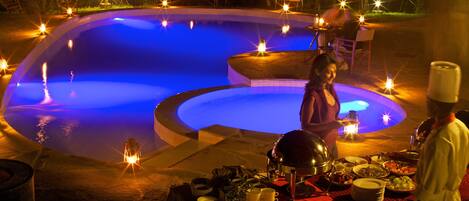 A poolside dining area with a table set for a meal, surrounded by candles and a chef in uniform.