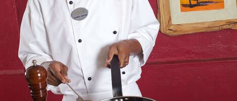 Un chef con uniforme blanco y un pañuelo rojo, sosteniendo una sartén con comida frente a una pared roja con un cuadro enmarcado.