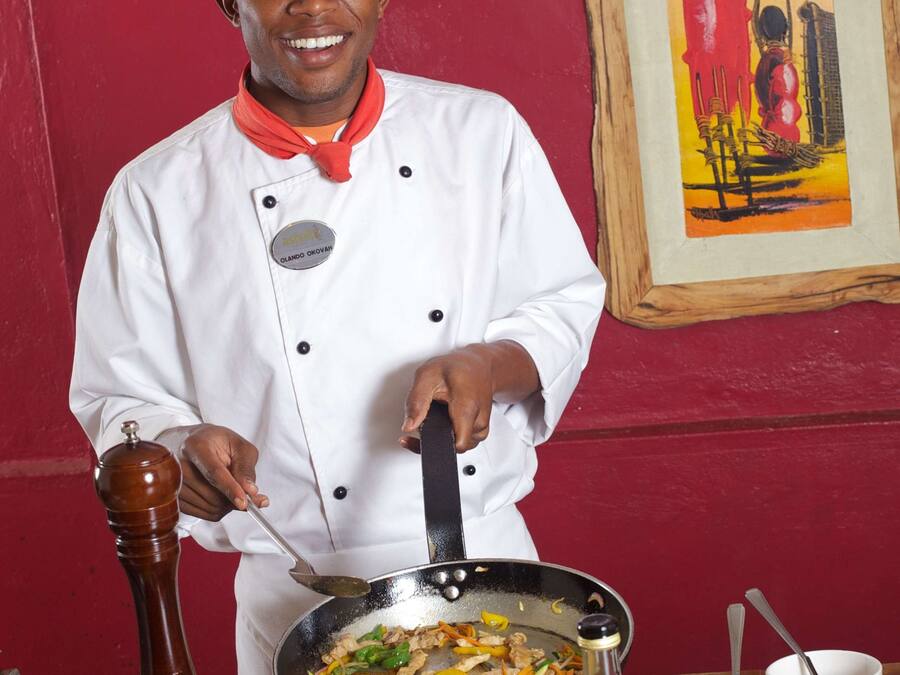 Un cuiner amb uniforme blanca i una faixa vermella, agafa una paella amb menjar, de peu davant d'una paret vermella amb un quadre encimbellat.