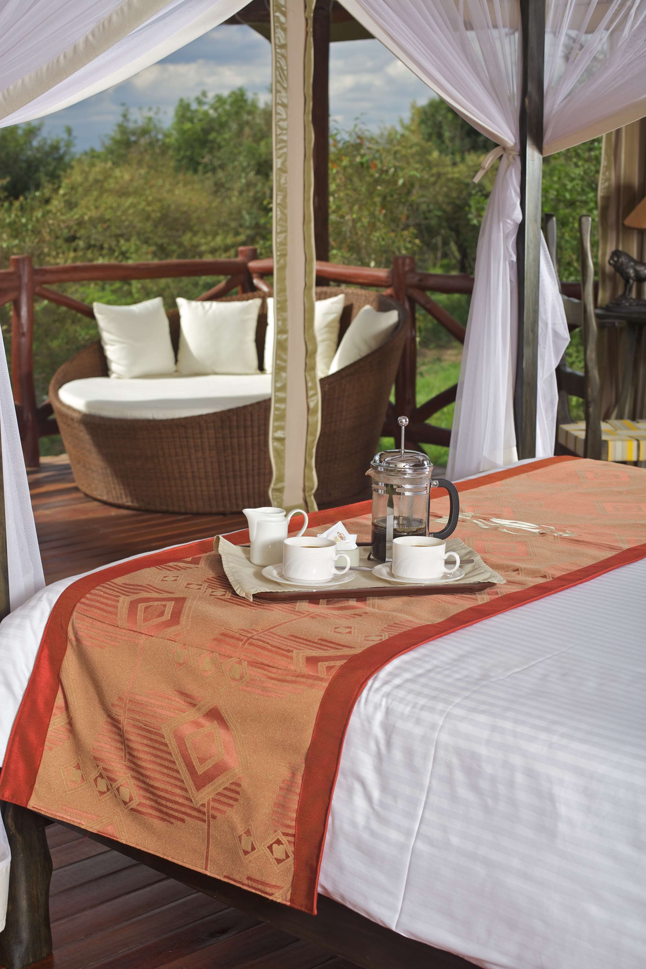 A bed with a tray of coffee and cups, a wicker chair with cushions, and a view of greenery.
