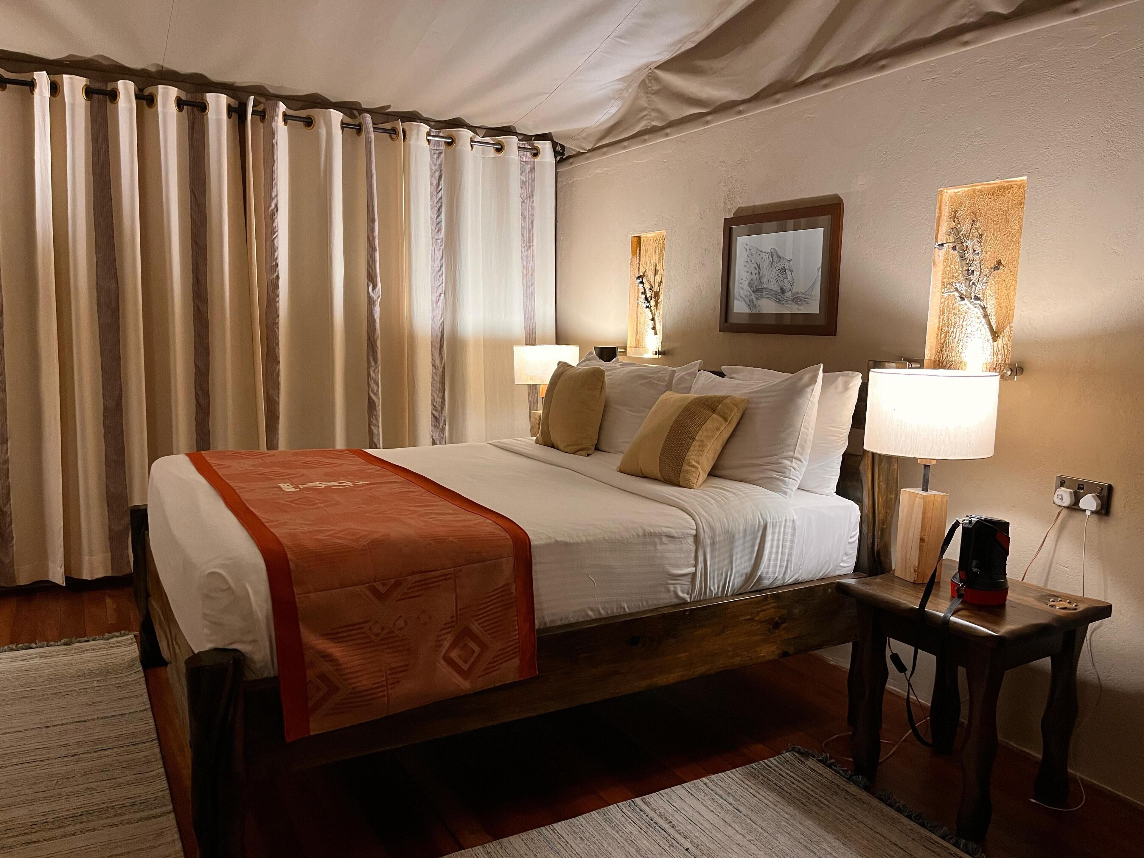 Habitación de hotel con cama de madera, cortinas color beige, dos lámparas de noche, una mesita de noche con lámpara y un cuadro en la pared.