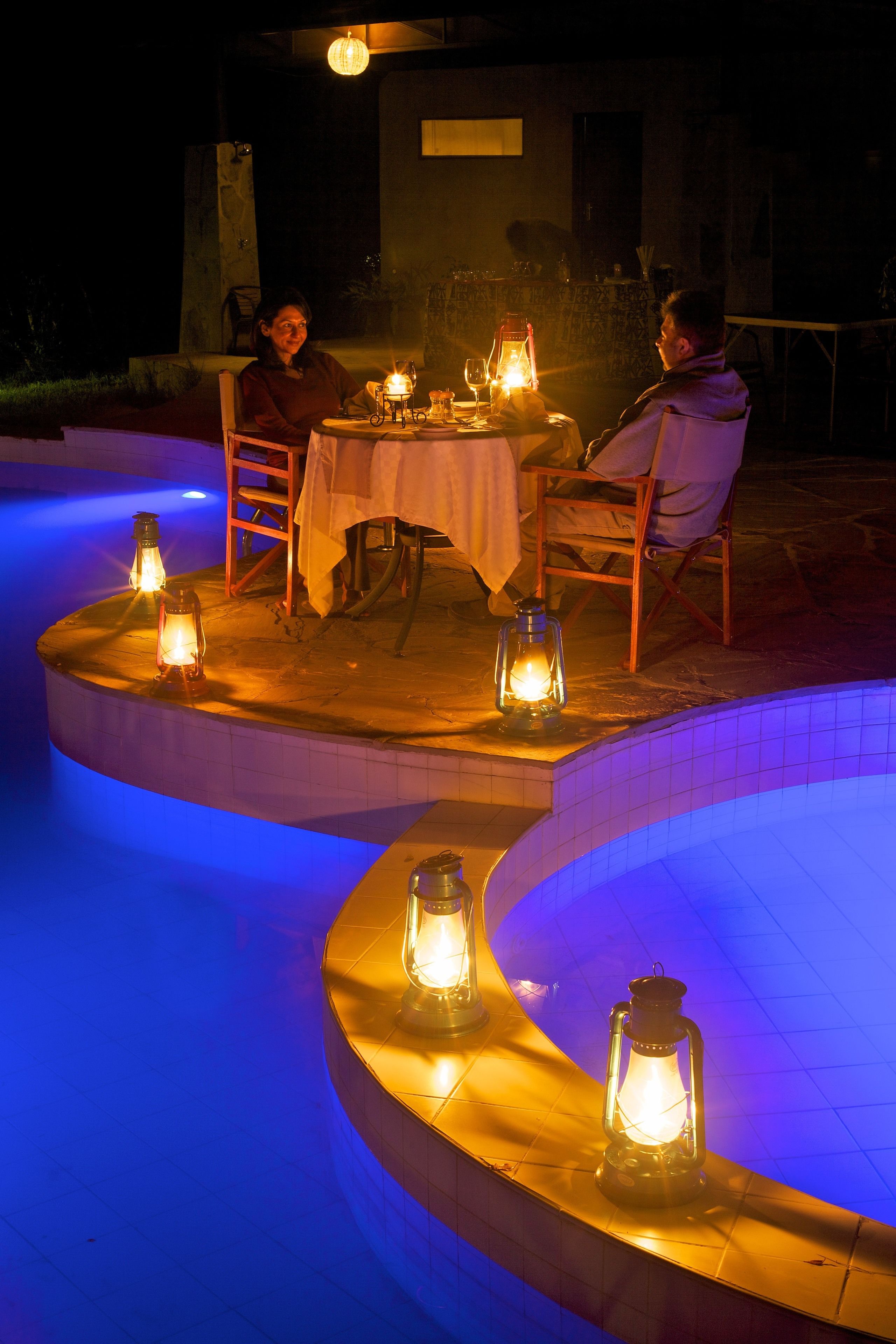 A poolside dining area with a table set for two, surrounded by lanterns.