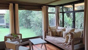 A room with a wooden floor, a sofa with cushions, a small table, and a chair. There are large windows with curtains, and a view of greenery outside. - Ashnil Mara Camp (Maasai Mara)