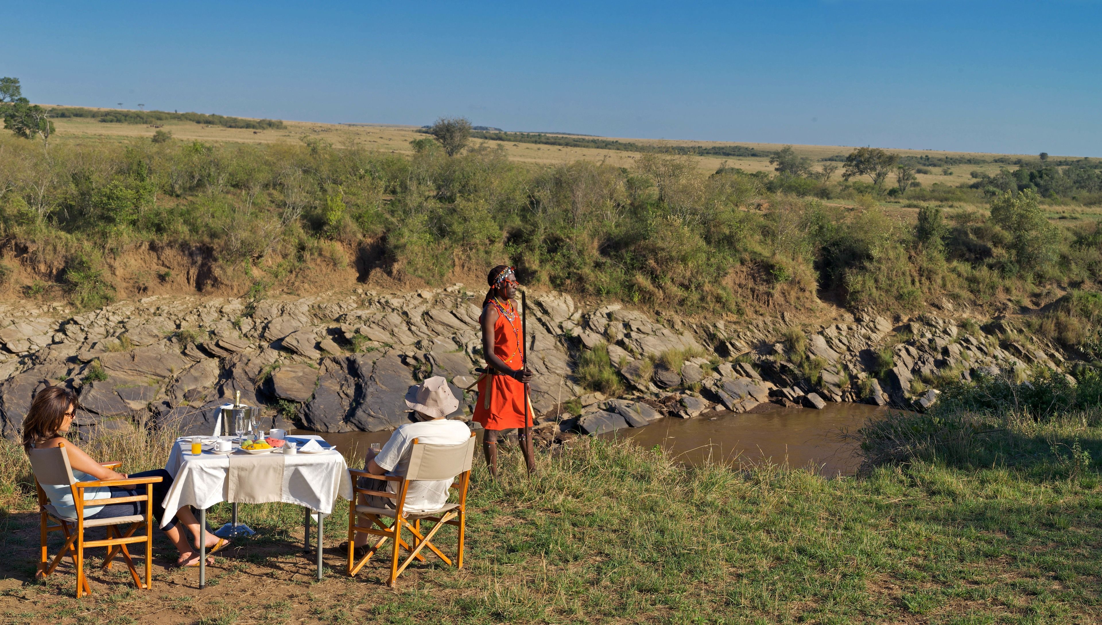 A table set up by a river with two people seated and a person standing nearby.