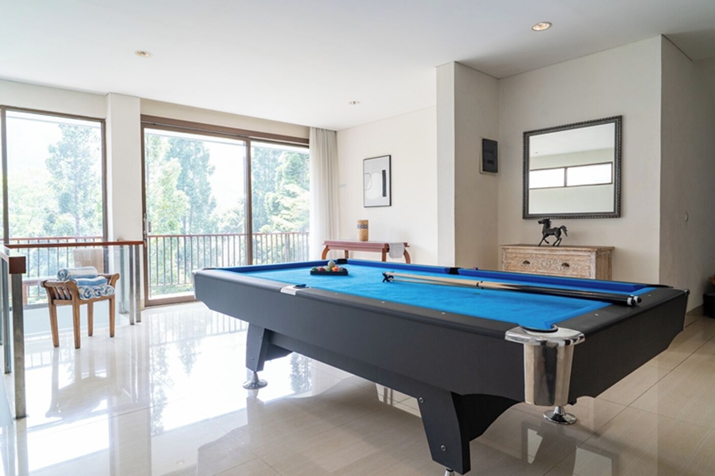 Una mesa de billar en una sala bien iluminada con ventanas amplias, una silla y una pequeña mesa con un adorno.