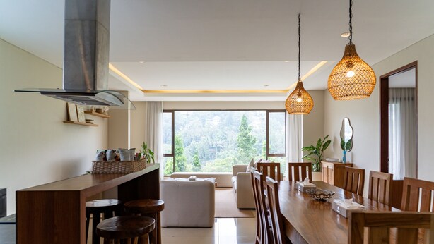 Una cocina moderna con una isla grande, mesa de comedor de madera y sillas. Se ve follaje a través de ventanas amplias.
